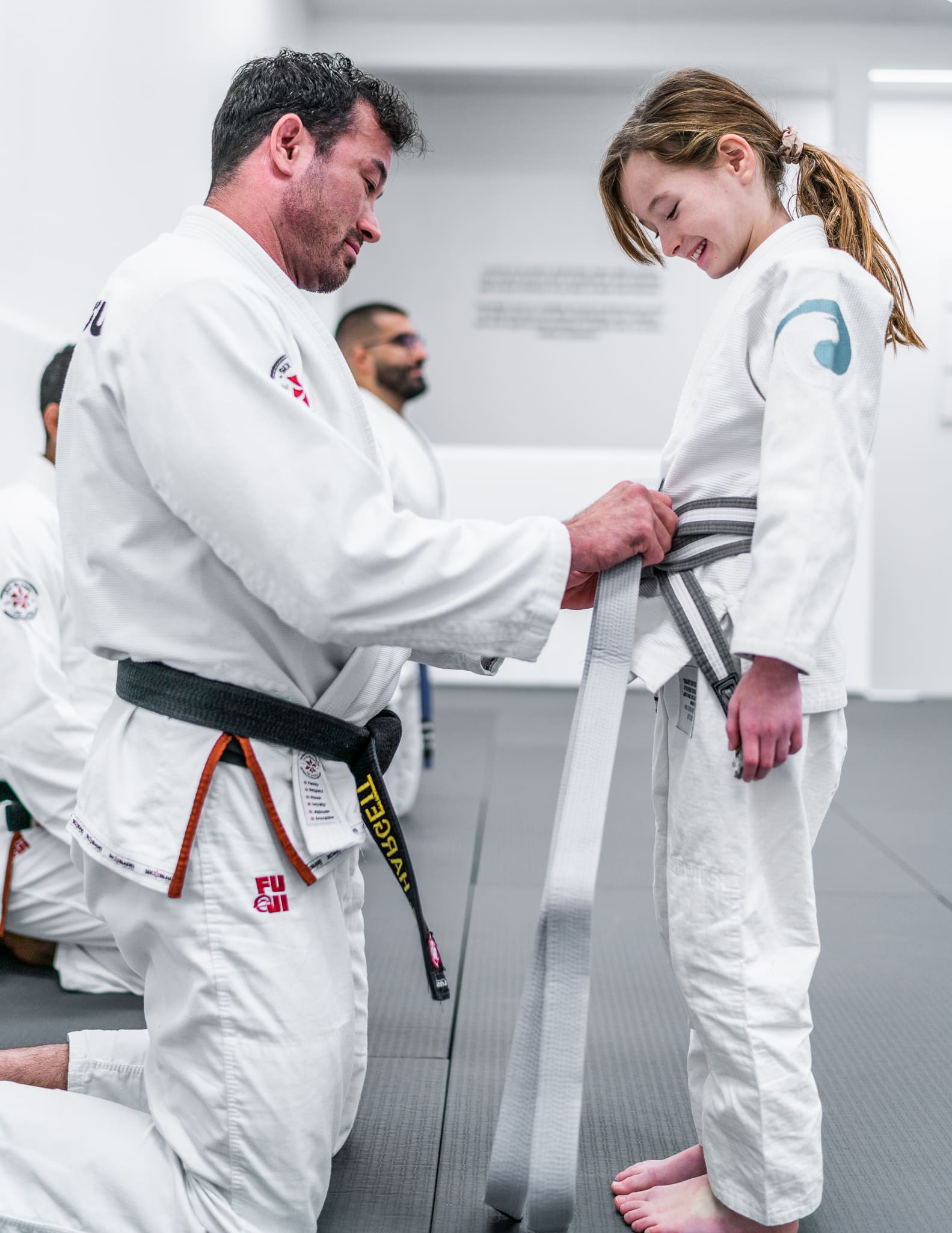A smiling Jiu-Jitsu instructor helping a young female student tie her new grey belt